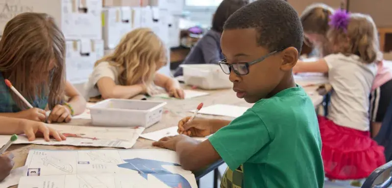 Écolier en pull vert écrivant sur une feuille blanche dans une salle de classe