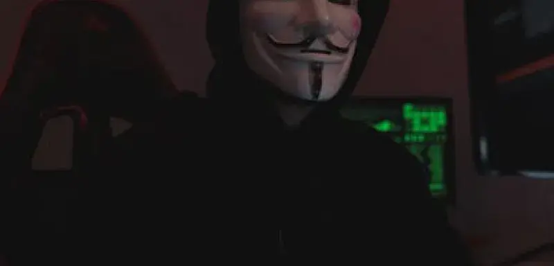 Silhouette mystérieuse portant un masque de Guy Fawkes, éclairée par des écrans d'ordinateur dans une pièce sombre