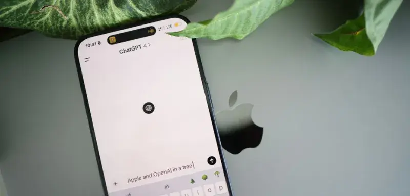 A smartphone shows a ChatGPT interface placed on an Apple laptop in a leafy environment.